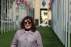 [ai] A woman wearing sunglasses and a purple coat stands on a grassy area surrounded by flagpoles displaying various international flags, with a building and a car visible in the background.